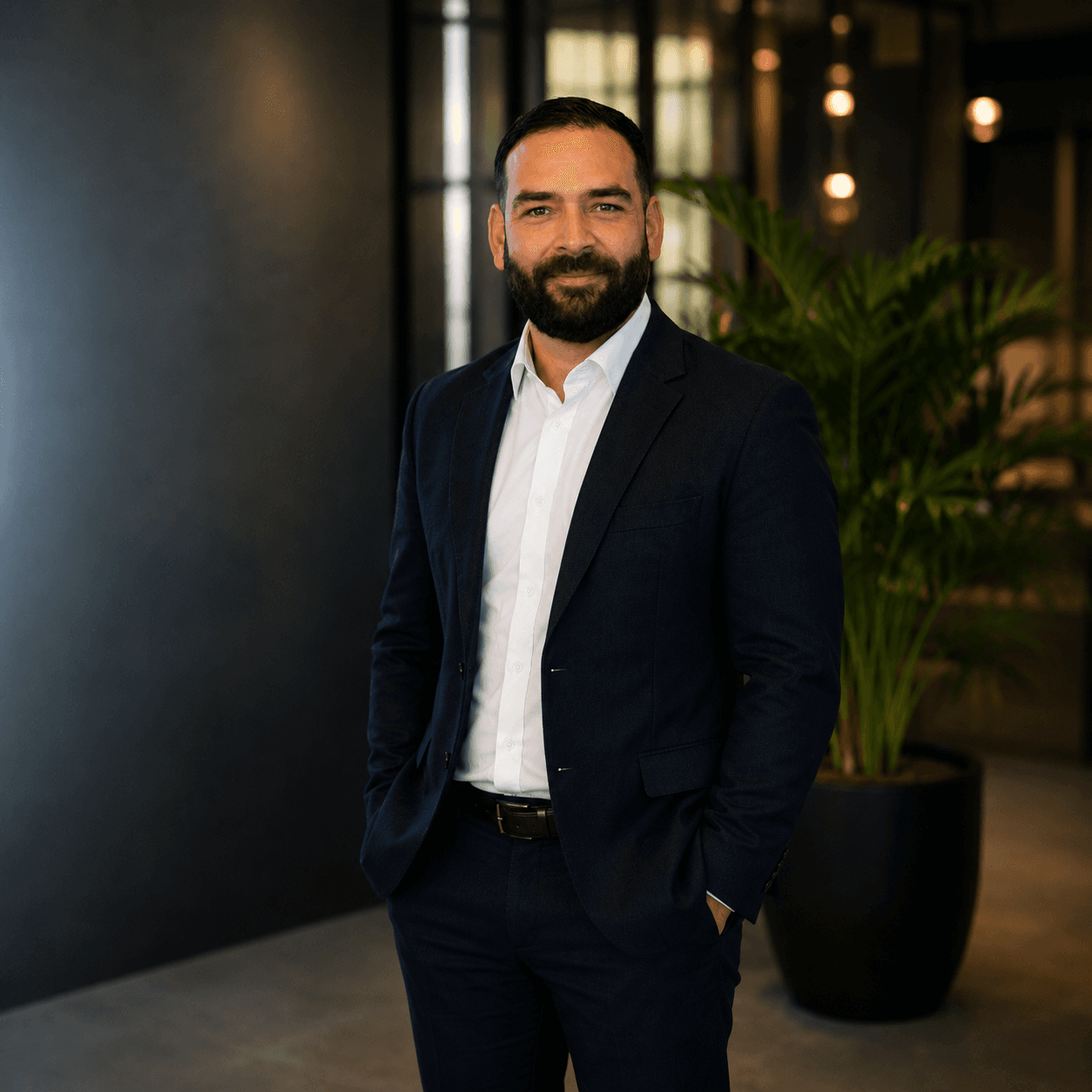 Bearded man in a navy suit and white shirt standing in a modern office.